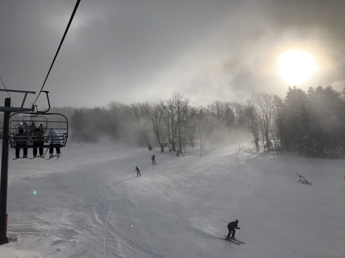 View of the ski slope below from a chairlift at Canaan Valley Ski Resort. In the image, there are skiers carving turns and the sun peaking out from above trees in the distance, cutting rays of sunlight through a snowy fog.