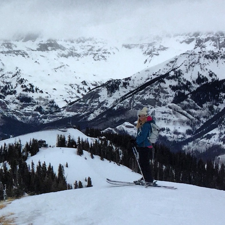 A woman skier in a blue coat, white hat, and a backpack overlooks a Telluride ski run backdropped by foggy snow-capped peaks. 
