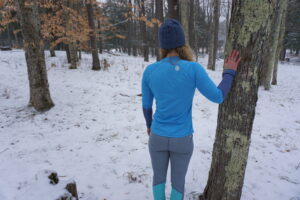 A woman facing away from the camera wearing gray baselayer bottoms and a blue baselayer shirt with a blue beanie stands to the left of a tree. Her right hand is touching the tree.