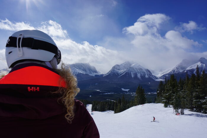 A woman in a red coat and a white helmet peers out at a snowy ski resort backdropped by snow-capped peaks against a cloud-speckled sky.