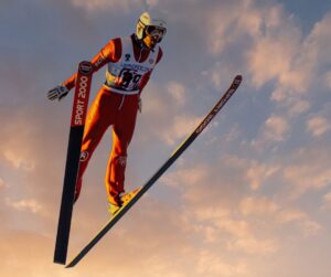 An Olympic freeskier in a red outfit and a helmet pictured mid-jump silhouetted against a pink sky with wispy clouds.