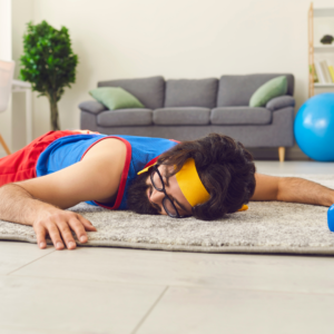Man lying face flat on carpet in a house. He has workout clothes on and a bar bell just out of reach of his left hand.