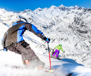 Backside view of two skiers carving turns on the edge of a descent into a steep bowl. 