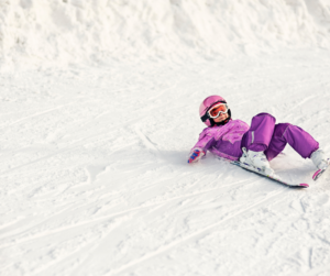 A girl in purple ski gear with white skis and boots on has just fallen down and is lying on her back in the snow with her hands out at her sides.