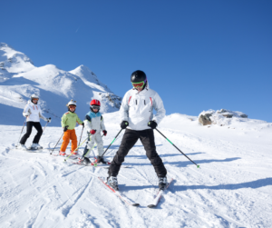 An adult skier guides three younger skiers down a gentle snowy slope backdropped by snow-capped peaks and a clear blue sky.