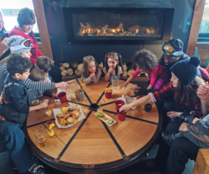 A family enjoys food and drinks at a round table by a fireplace after a day of skiing.