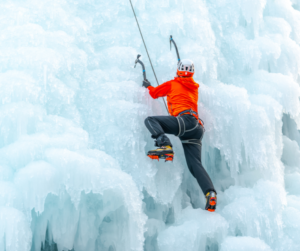 A person in a red coat on top rope is ice climbing using ice axes and crampons up a gradually sloping ice feature.