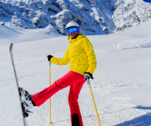 A skier dressed in a bright yellow jacket and neon pants poses for the camera with one left lifted in the air. 