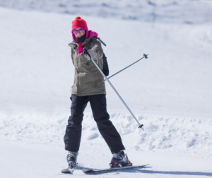 A skier in the pizza stance coming down the slopes looking at the camera. She has her left hand holding a ski pole raised in a thumbs up position. 