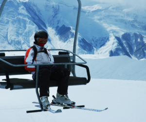 A skier in a white jacket with red stripes rides solo on a chairlift backdropped by snowy peaks. 