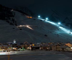 View of a snowy ski resort village lit up at night at the base of an illuminated ski slope. 