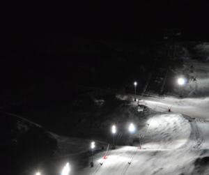 A ski resort lit up at night for night skiing. Lights run the length of a chairlift operating over a groomed slope. 