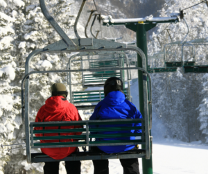 View from behind of two people on a ski resort chairlift riding through a wintry wonderland. 
