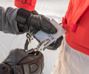 Picture of a person with black gloves punching a hole into a skier's lift ticket hanging from a red jacket. 