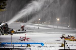 Two snow machines blowing out snow under lights at nighttime at a ski resort. 