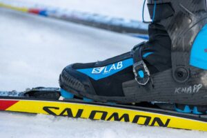 Close up left side view of a blue and black ski boot strapped into ski bindings on a yellow Salomon-labeled ski. 