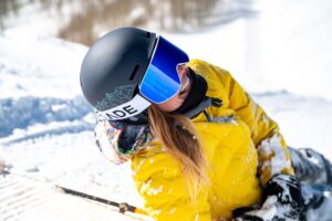 Woman in a yellow jacket with ski goggles over a black helmet posed sideways in the snow.