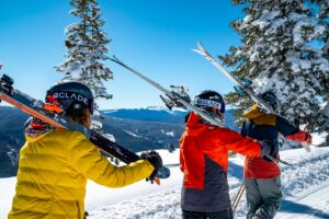 Three skiers carrying skis on their shoulders backdropped by snowy trees during a group ski lesson.