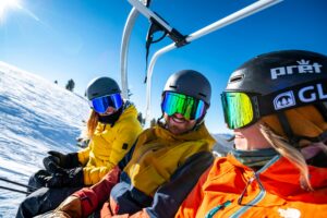 Three friends on a ski lift are chatting with each other. They're all wearing helmets and ski goggles. 