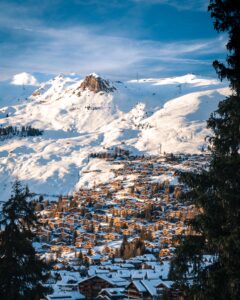 View of a sprawling ski town backdropped by snowy mountain peaks from a distance. The frame features two outlines of trees in the foreground. 